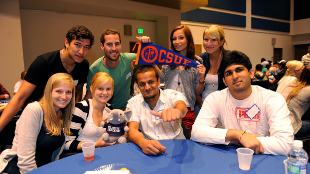 A group of German and Indian students gather around a table and wave a CSUF flag.