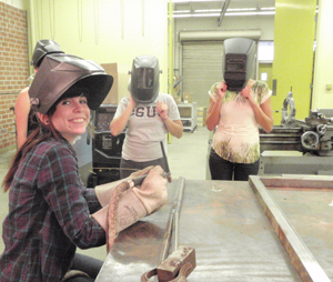 Three women wearing welding helmets in one of the engineering labs.