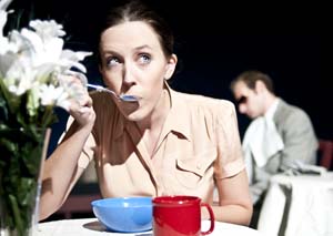 A young woman sips soup while in the background an actor protrays a dean man.