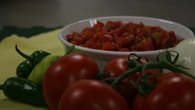 A bowl of salsa surrounded by peppers and tomatoes on a yellow cloth