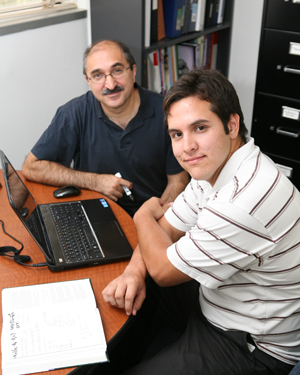 Two men sit at a table looking at a laptop computer.