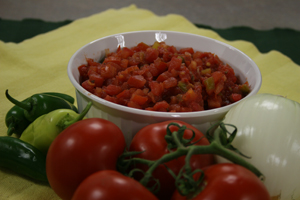 bowl of salsa surrounded by red tomatoes, green chilies and a white onion on a yellow cloth napkin.