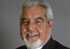 close up photo of a white haired and bearded man in a black suit against a gray background.