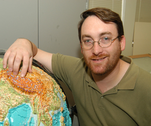 David Bowman in a green shirt and standing next to a globe of the world.