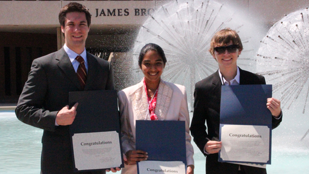 Two male students on either side of a female student — all standing in front of a fountain 