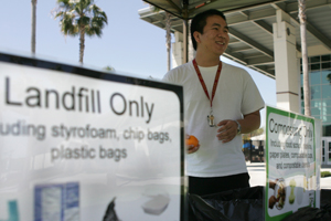 A young man mans a station with three trash barrels marked landfill, composted and recycle.
