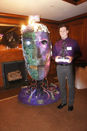 Young man stands next to his winning sculpture of a giant head made of various electronic materials.