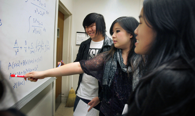 Three students look at a white board covered with mathematical problems.