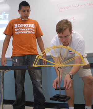 Two male students set a weight dangling from their spaghetti bridge.