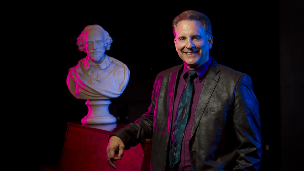 Jim Volz, in black suit and fushia shirt, stands beside an upright piano and a bust of William Shakespeare.