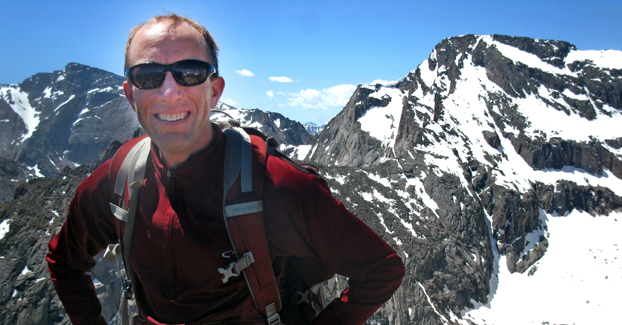 Man wearing red jacket and hiking gear stands before a range of snow-capped mountains.