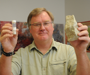 Jeffrey Knott holding a chunk of volcanic rock in one hand and a vial of crush rock in the other.