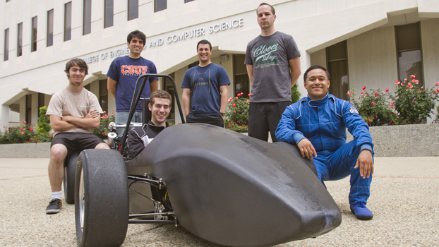 five men surround a small race car in which a man sits in the drivers seat.