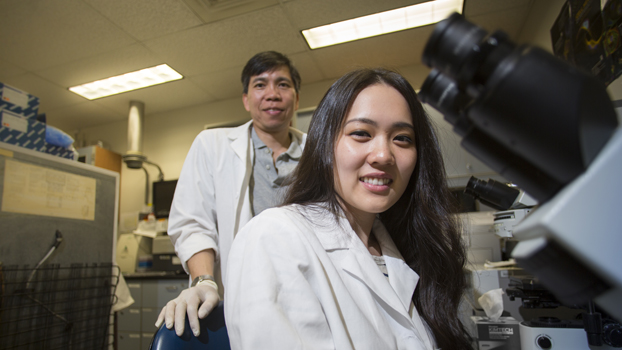 Faculty member stands behind a student using a microscope.